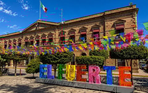 Historic yellow stone building with Mexican flag, colorful WELCOME sign in foreground, and vibrant paper banners hanging across facade