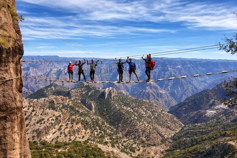 Group of people walking on a narrow cable suspension bridge high above a vast canyon landscape with mountains and valleys below