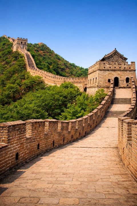 Ancient Great Wall of China with stone watchtowers, fortified walls winding through green mountainous terrain under blue sky