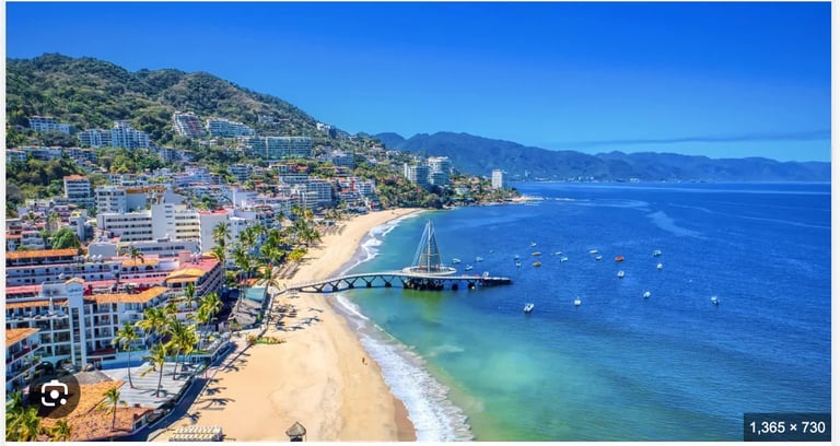 Aerial view of a Mediterranean coastal resort town with sandy beach, sailboat, turquoise water, and hillside buildings under clear blue sky
