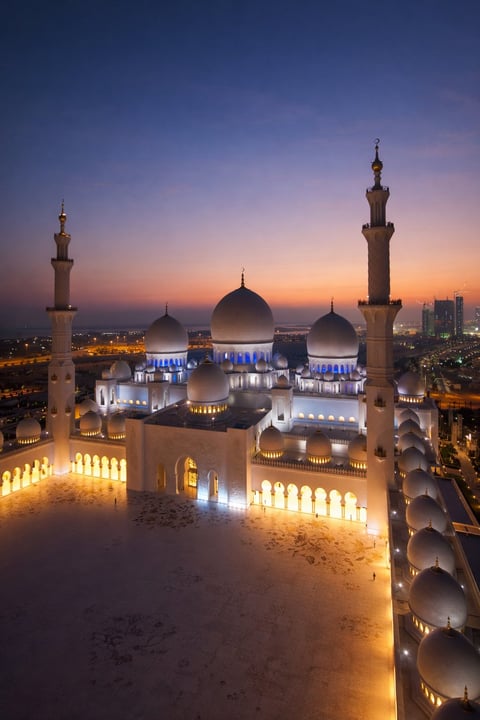 Grand mosque at dusk with illuminated domes, minarets, and courtyard at twilight, city skyline in background