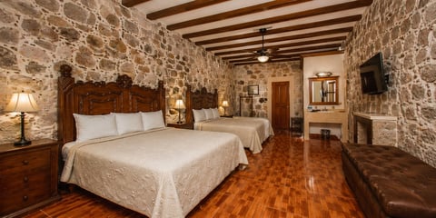 Rustic hotel bedroom with two beds, exposed stone walls, wooden beams, hardwood floors, and period furniture