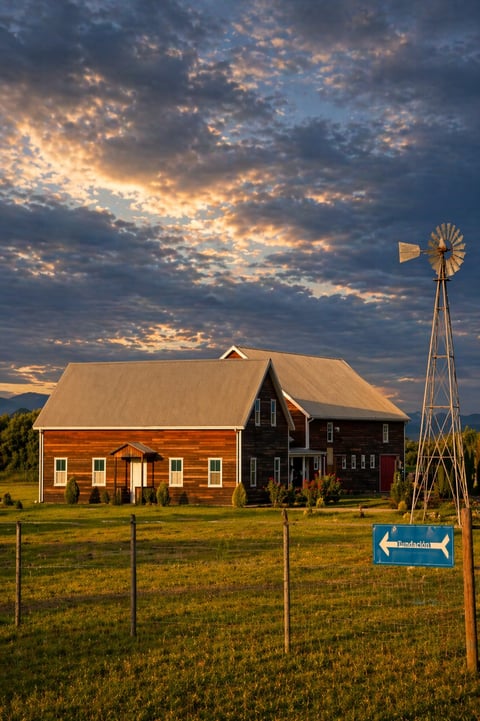 Red barn with windmill under dramatic sunset sky, fenced pasture with directional sign in rural landscape