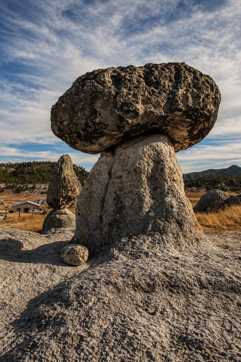 Large balanced rock formation with a precariously perched boulder on top, set in a desert landscape with mountains in the background