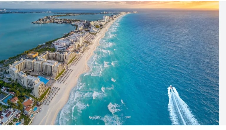 Aerial view of a tropical beach resort with turquoise waters, sandy coastline, high-rise hotels, and a speedboat creating a wake in the ocean at sunset.