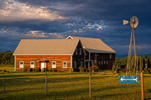 A red barn with tan roof and metal windmill stand behind a wooden fence in a rural prairie landscape at dusk