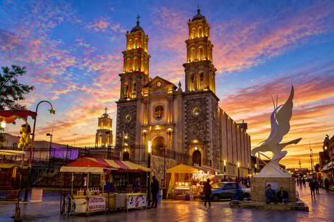 Illuminated cathedral with twin domes at sunset in a plaza with street vendors and a sculpture, vibrant orange and purple sky